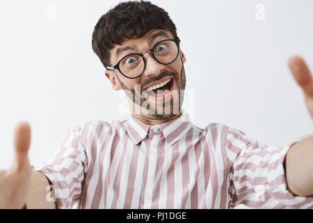 Nahaufnahme der lustig, gefühlvoll und glücklich, gut aussehende moderne männliche Hipster in Gläsern und Rosa gestreifte Shirt ziehen die Hände in Richtung Kamera selfie, äfft zu machen und Grimassen, um täuschen Stockfoto