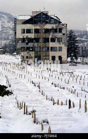 4. März 1993 während der Belagerung von Sarajevo: ein stark beschädigtes Gebäude steht unten Reihen der schneebedeckten bosnisch-muslimische Gräber in der Lion Friedhof, direkt unter den Kosevo Krankenhaus. Die Gräber sind alle 1992. Stockfoto