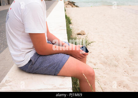 Ansicht der männlichen Kaukasier Teenager mit weißem T-Shirt und Jeans Shorts sitzen auf einer Wand, die mit dem Strand und Meer sein Smartphone hält in seinen Händen 7/8 Stockfoto