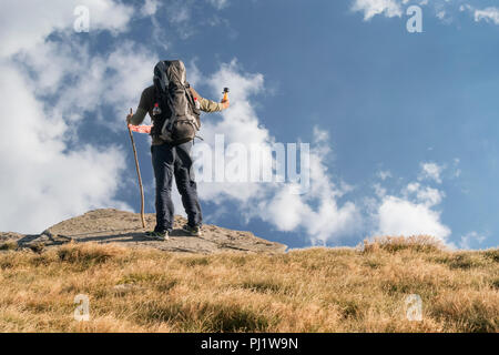 Touristen mit einem großen Rucksack auf dem Rücken steht auf dem Gipfel des Berges, hält eine Kamera in die Hand und nimmt Bilder des Himmels Stockfoto