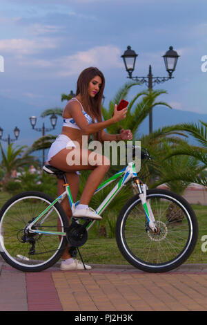 Schöne langhaarige junge Frau mit dem Fahrrad Spaß hat, mit einem Smartphone, macht selfy auf einem Smartphone Stockfoto