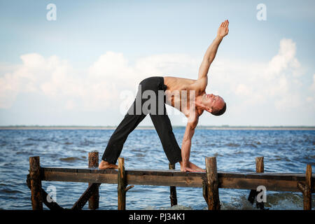 Junge yoga Trainer üben parivrita trikonasana oder Dreieck Pose auf einer hölzernen Pier an einem See oder Fluss Ufer. Gesunder Lebensstil Konzept Stockfoto