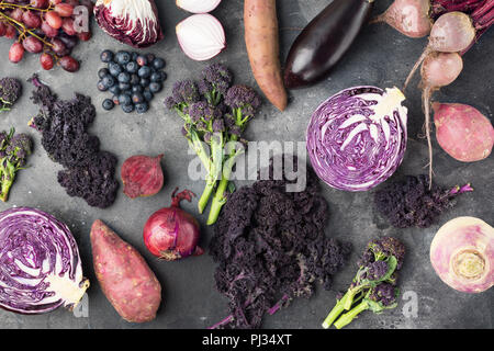 Gemüse und Obst Hintergrund in lila, grün und Dunkelrot Farben, Rotkohl Grünkohl Rosenkohl Kartoffel Zwiebeln Brokkoli Heidelbeeren Trauben, Ansicht von oben, dunkelgrauer Hintergrund, selektiver Fokus Stockfoto