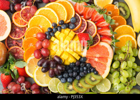 Gesunde Früchte Hintergrund in Regenbogenfarben Orangen, Äpfel, Trauben, Birnen, Mango, Erdbeeren, Kiwis auf dem grauen Beton Tabelle, Ansicht von oben, selektiver Fokus Stockfoto