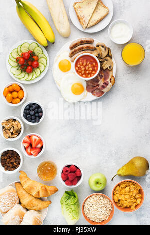Gesundes Frühstück gebratene Eier, Würstchen, Speck und Pilzen mit Auswahl an Obst und Gemüse, Brot und Saft auf das grau weiß Tisch, top vie Stockfoto