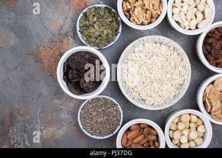 Zutaten für hausgemachtes Müsli, Hafer, verschiedene Nüsse und Rosinen, Datteln, Samen auf dem grauen Stein Hintergrund, Ansicht von oben Stockfoto