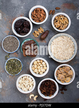 Hausgemachtes Müsli Zutaten, Hafer, verschiedene Nüsse und Rosinen, Datteln, Samen auf dem grauen Stein Hintergrund, Ansicht von oben Stockfoto