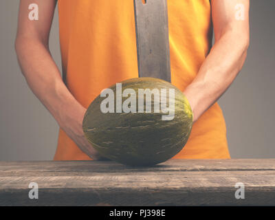 Junger Mann mit einem alten Cleaver und eine frische Melone auf eine rustikale Küche Tabelle Stockfoto