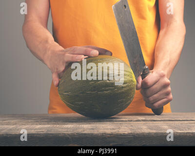 Junger Mann mit einem alten Cleaver und eine frische Melone auf eine rustikale Küche Tabelle Stockfoto