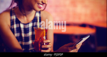 Junge Frau mit Mobiltelefon, während Sie einen Cocktail trinken Stockfoto