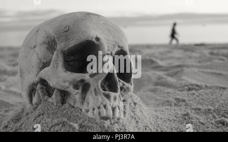 Menschlichen Kopf Schädel halb im Sand begraben mit menschlichen Wandern in der Hintergrund verschwommen Stockfoto