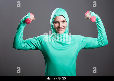 Gerne schöne muslimische Frau in Grün hijab oder islamischen Sportswear, die hielten rote Kurzhanteln, erhobenen Armen, an der Kamera und toothy Lächeln suchen. Stockfoto