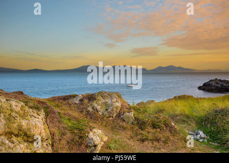 Sonnenuntergangsseelandschaft von Ynys Llanddwyn Headland (Gezeiteninsel in Anglesey, Wales) über Meer in Richtung Yr Eifl (die Rivalen auf Englisch) & entferntes Boot. Stockfoto