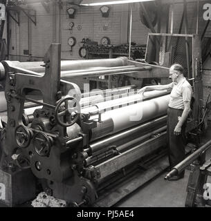 1950, historische, am Brittains Papierfabrik, ein männlicher Arbeiter beaufsichtigt ein großes Papier Presse Maschine mit großen stahlrollen oder 'Kalanderwalzen" ein Papier mit einer glatten Oberfläche, England, UK zu produzieren. Stockfoto