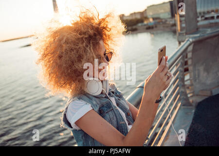 Trendige Mädchen mit großen Kopfhörer und Sonnenbrille auf einer Stadt spazieren, junge Frau mit einem Smartphone. Portrait einer jungen hübschen Blondine mit üppigen Locken, m Stockfoto