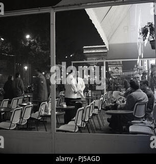 1950er Jahre, abends mal in Paris, Frankreich und zwei Kellner zusammen chatten auf dem Bürgersteig Terrasse eines Cafés auf den Champs Elysées, nur wenige Schritte von der Place de Trioumphe Denkmal, die größte und berühmteste Arc in der Welt. Stockfoto