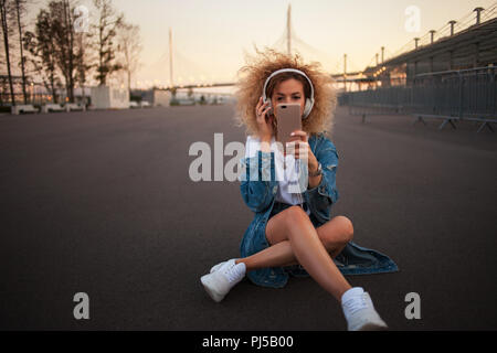 Glückliche junge Mädchen auf der Straße ein Foto auf einem Smartphone. Schöne Blondine mit Kopfhörern und Smartphone ein selfie machen. im Freien Stockfoto