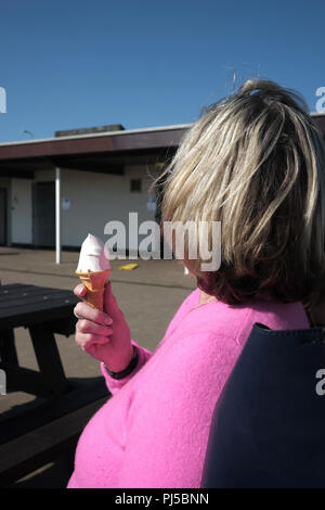 Rückansicht, siebzig Jahre alte britische Frau mit rosa Kaschmir top in eine Pause von ihrem Vanilleeis Essen mit Konus am Meer, Stockfoto