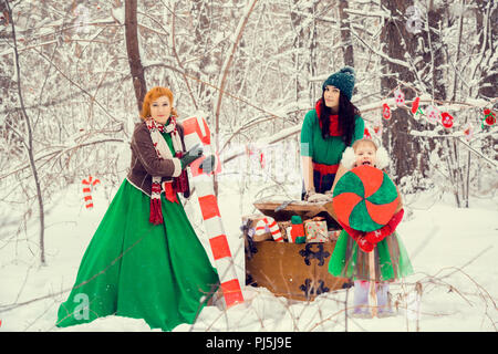 Familie in der traditionellen Weihnachtsstern Kostüme, rot-grüne, Helfer Elfen Santa's, im Winter Wald mit einer Truhe von Geschenken und einem riesigen Candy Stockfoto