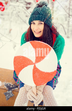 Brunette Mädchen in einem grünen Hut und in ein Kostüm von Blumen traditionellen zu den elfen Santa's Helfer im Winter Forest unter dem Schnee mit einer Truhe, der Geschenke und der riesigen Candy Stockfoto