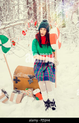 Brunette Mädchen in einem grünen Hut und in ein Kostüm von Blumen traditionellen zu den elfen Santa's Helfer im Winter Forest unter dem Schnee mit einer Truhe, der Geschenke und der riesigen Candy Stockfoto