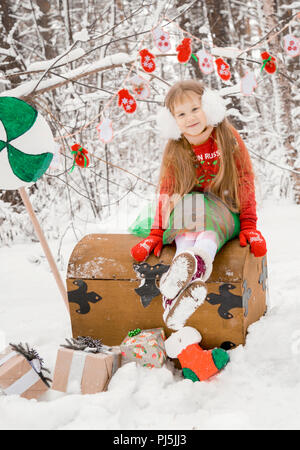 Eine kleine Rothaarige Mädchen mit langen Haaren in einem üppigen Rock und einem grünen Mantel mit Spitze, in das Kostüm von Zwerg assistant Santa Claus im Winter Wald mit riesigen süssigkeit, eine Truhe von Geschenken in einem Korb Weihnachtskugeln sammelt Stockfoto