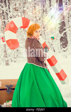 Feurige rothaarige Frau in einer Kugel grünes Kleid mit einem roten Ledergürtel in das Kostüm von Zwerg assistant Santa Claus im Winter Wald mit riesigen Süßigkeiten, Geschenke, das Konzept des neuen Jahres Stockfoto