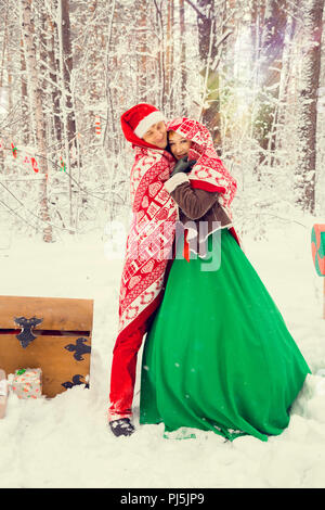 Junges Paar, Mann und Frau, Mann und Frau sind zu Fuß in Kostümen von Blumen, die typisch für die Elfen von Santa's Helfer in einem Winter Forest unter dem Schnee mit einer Truhe voller Geschenke und ein riesiges Candy Stockfoto