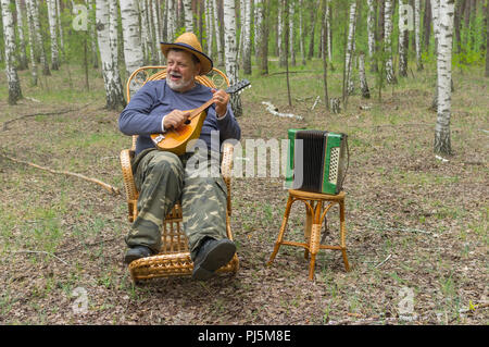 Senior singen und spielen Mandoline, während in einem wicker Schaukelstühlen in Birke Wald sitzen Stockfoto