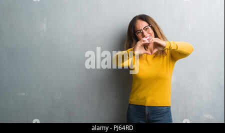 Mittleres Alter spanischer Frau über graue Wand Brillen in der Liebe mit Herz und mit Hand lächelnd. Romantische Konzept. Stockfoto