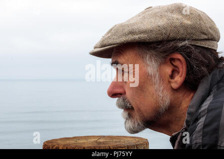 Schließen Sie sich an Profil von nachdenklich, ernst älterer Mann mit einem grauen Bart tragen eines Tweed Cap in die Ferne mit Ozean Horizont im Hintergrund suchen Stockfoto