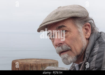 Nahaufnahme von nachdenklich, ernst älterer Mann mit einem grauen Bart tragen eines Tweed Cap an Kamera suchen mit Ozean Horizont im Hintergrund Stockfoto