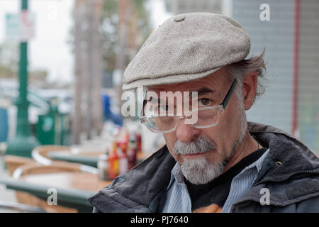 Schließen Sie sich an Profil von nachdenklich, ernst älterer Mann mit einem grauen Bart trägt eine Kappe an der Kamera über Lesebrille, Bokeh verschwimmt Outdoor resta Stockfoto