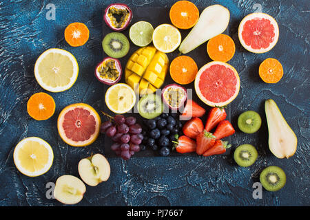 Vielfalt von bunten Früchten, Orangen, Zitronen, Kiwis, Grapefruits, Erdbeeren, Trauben, Heidelbeeren, Birnen, Passionsfrucht, Mango am schwarzen Schiefer Stockfoto