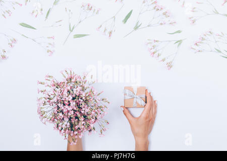 Woman's Hände, die kleinen Geschenkkarton mit Silber und Bogen Blumenstrauß unter Rosa Blumenmuster Grenze Rahmen auf weißem Hintergrund, flach. Ansicht von oben Mädchen mit einem Pr Stockfoto