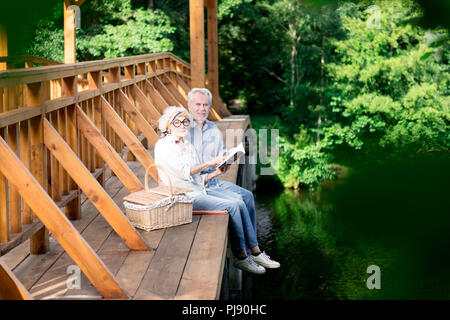 Im Alter von Paar lesen interessante Buch beim Sitzen auf der Brücke Stockfoto
