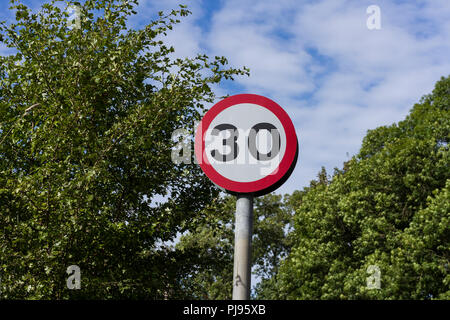 30 mph, rund, rot & weiß Britischen Straßenverkehr Zeichen gegen Bäume & blauer Himmel mit Wolken Hintergrund, England, Großbritannien Stockfoto