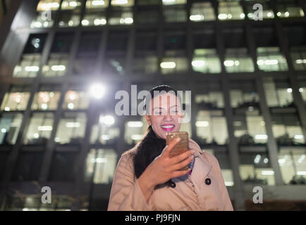 Lächelnd Geschäftsfrau mit smart phone unter städtischen Hochhaus in der Nacht Stockfoto
