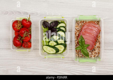 Kunststoff Mahlzeit prep Container mit frischen Erdbeeren, Gurken und Salat, gekochten Buchweizen Brei und Scheiben von Fleisch auf grau Holz- Hintergrund. Top Stockfoto