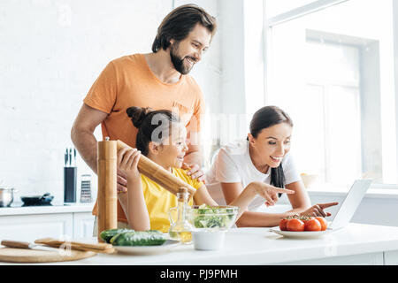 Schöne junge Familie mit Laptop, beim Kochen in der Küche Stockfoto