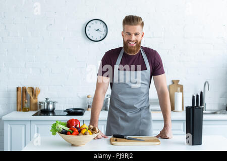 Gut aussehender bärtiger junger Mann in Schürze lächelte Kamera beim Kochen in der Küche Stockfoto