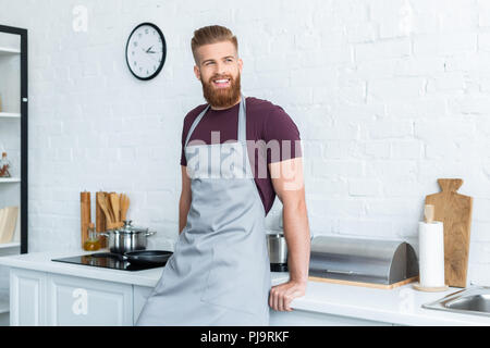 Gut aussehender bärtiger junger Mann in Schürze lächelnd und Wegsehen in der Küche Stockfoto