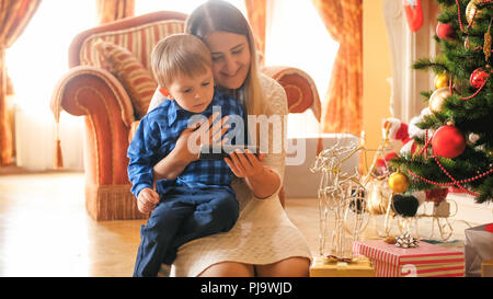 Portrait von Toddler boy wathcing Video auf Handy mit Mutter im Wohnzimmer für Weihnachten dekoriert Stockfoto