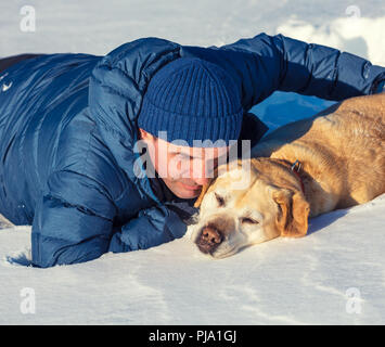 Ein Mann mit einem Labrador Retriever Hund liegend im Schnee im Winter. Stockfoto