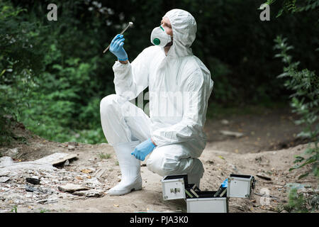 Fokussierte männliche Wissenschaftler in Schutzanzug und Maske Prüfung Stein beim Halten von Pinzette im Wald Stockfoto