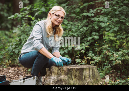 Lächelnd weibliche Wissenschaftler in Latex Handschuhe sitzen mit Pinzette und Lupe in Wald Stockfoto