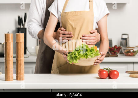 Zugeschnittenes Bild des jungen Paares umarmen und halten Schüssel mit Salat in der Küche Stockfoto