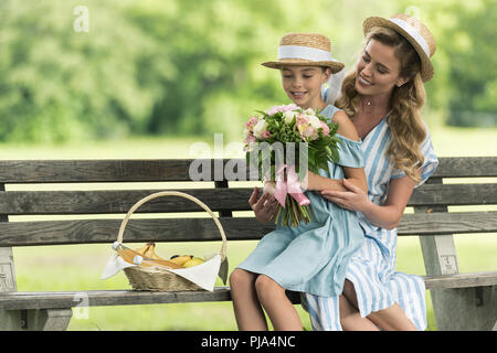 Lächelnde Mutter und Tochter mit Früchten in Weidenkorb und floralen Bouquet sitzt auf der Bank Stockfoto