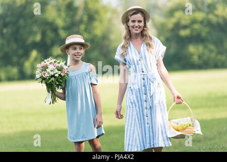 Lächelnd schöne Mutter und Tochter mit Früchten in Weidenkorb und Blumenstrauß auf dem grünen Rasen Stockfoto