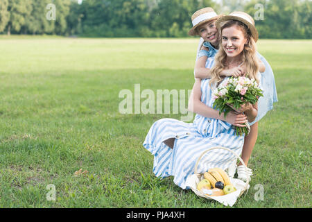 Schönen Mutter und Tochter mit Früchten in Weidenkorb und Blumenstrauß sitzen auf grünen Rasen Stockfoto